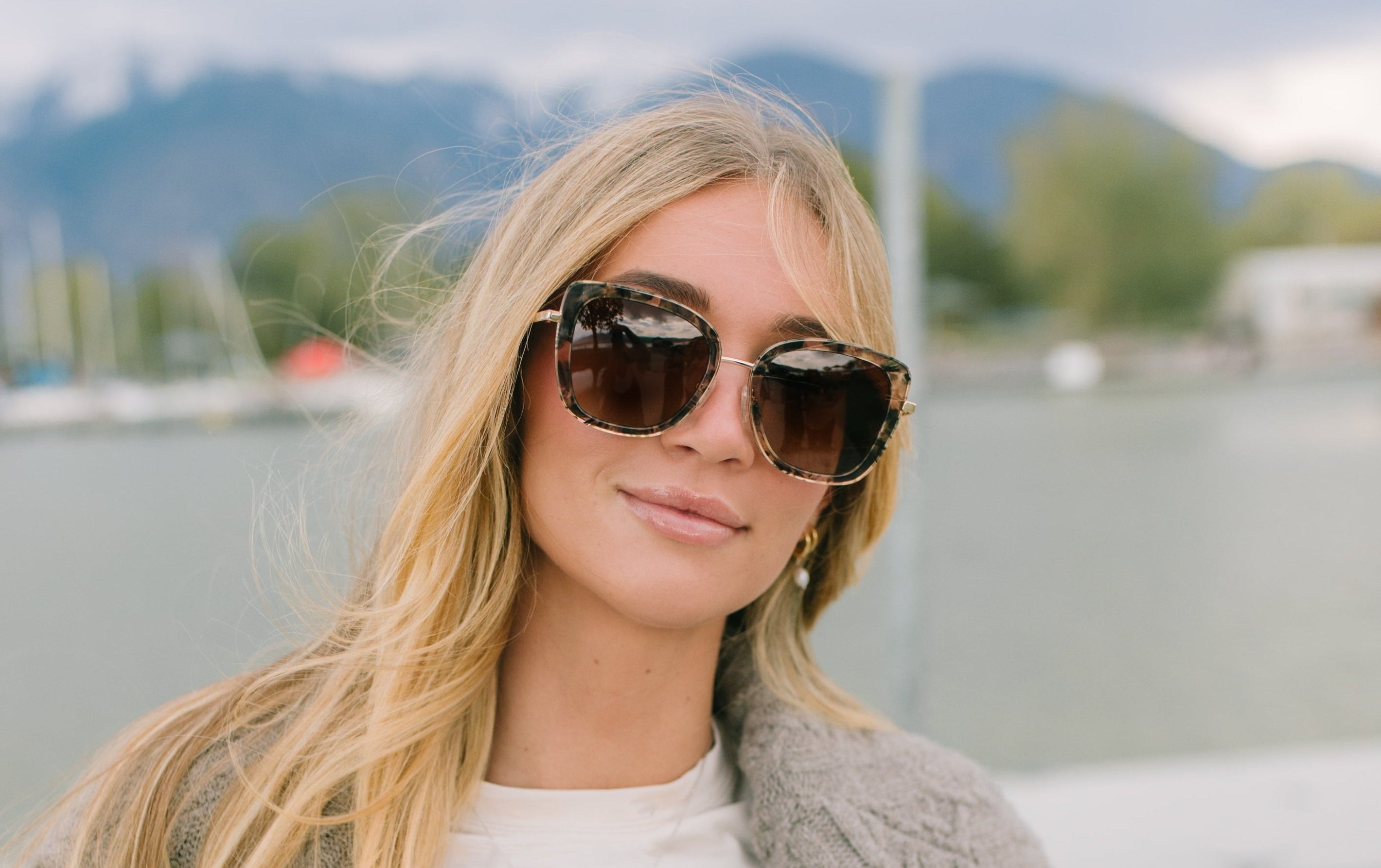 Woman wearing sunglasses and a gray cardigan by a waterfront with mountains in the background