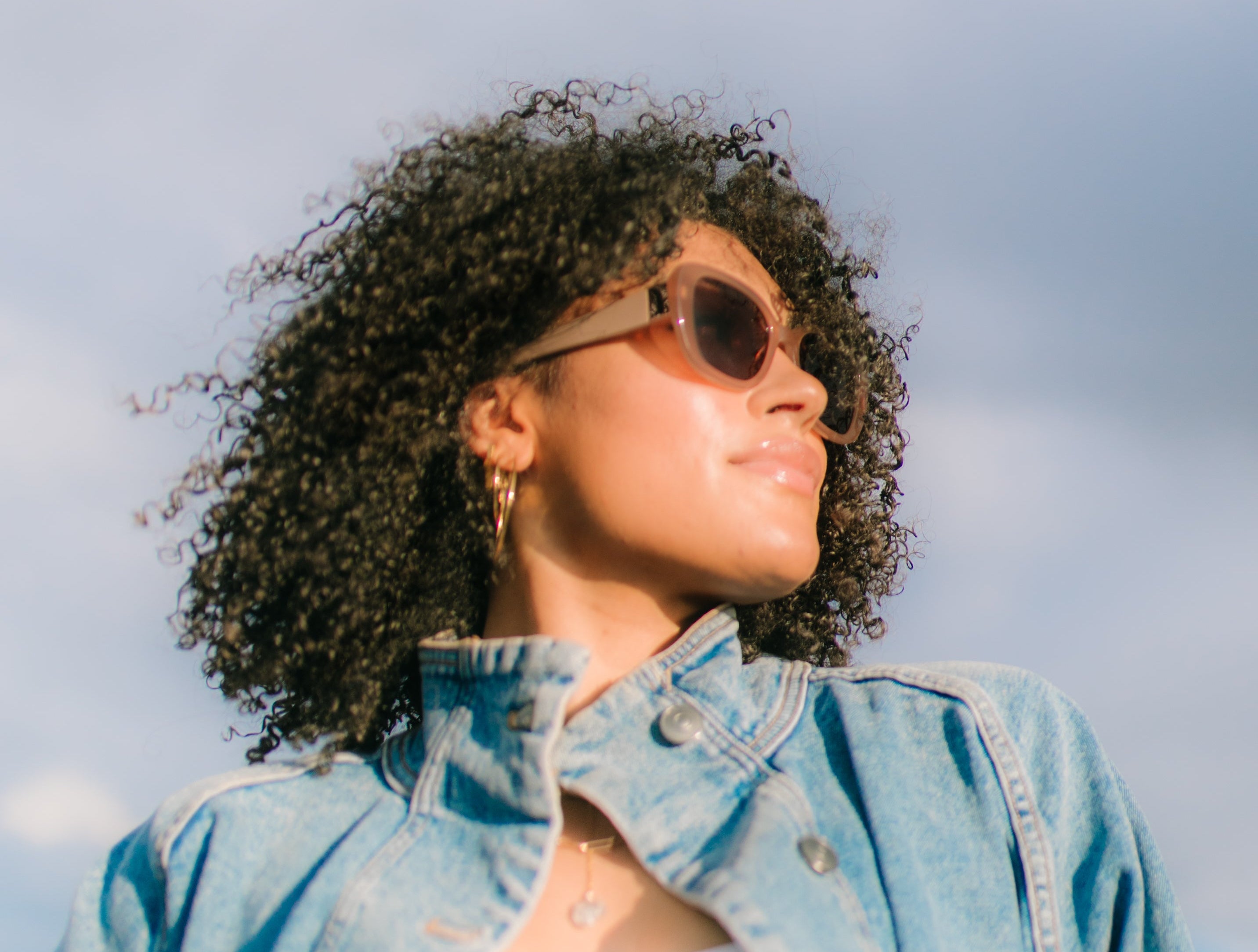 Woman wearing sunglasses and a denim jacket against a cloudy sky