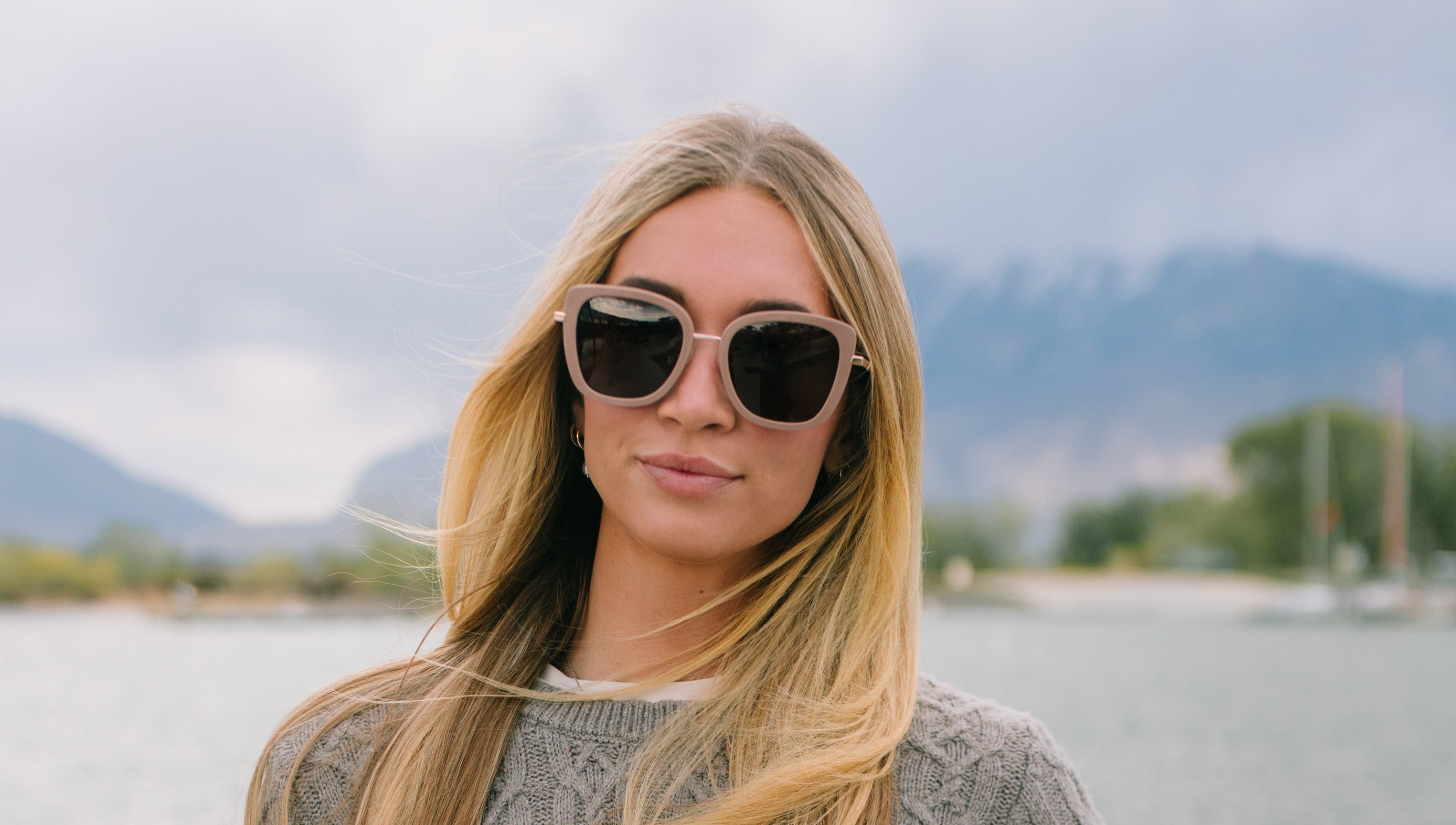 Woman wearing a gray sweater and sunglasses by a body of water with mountains in the background