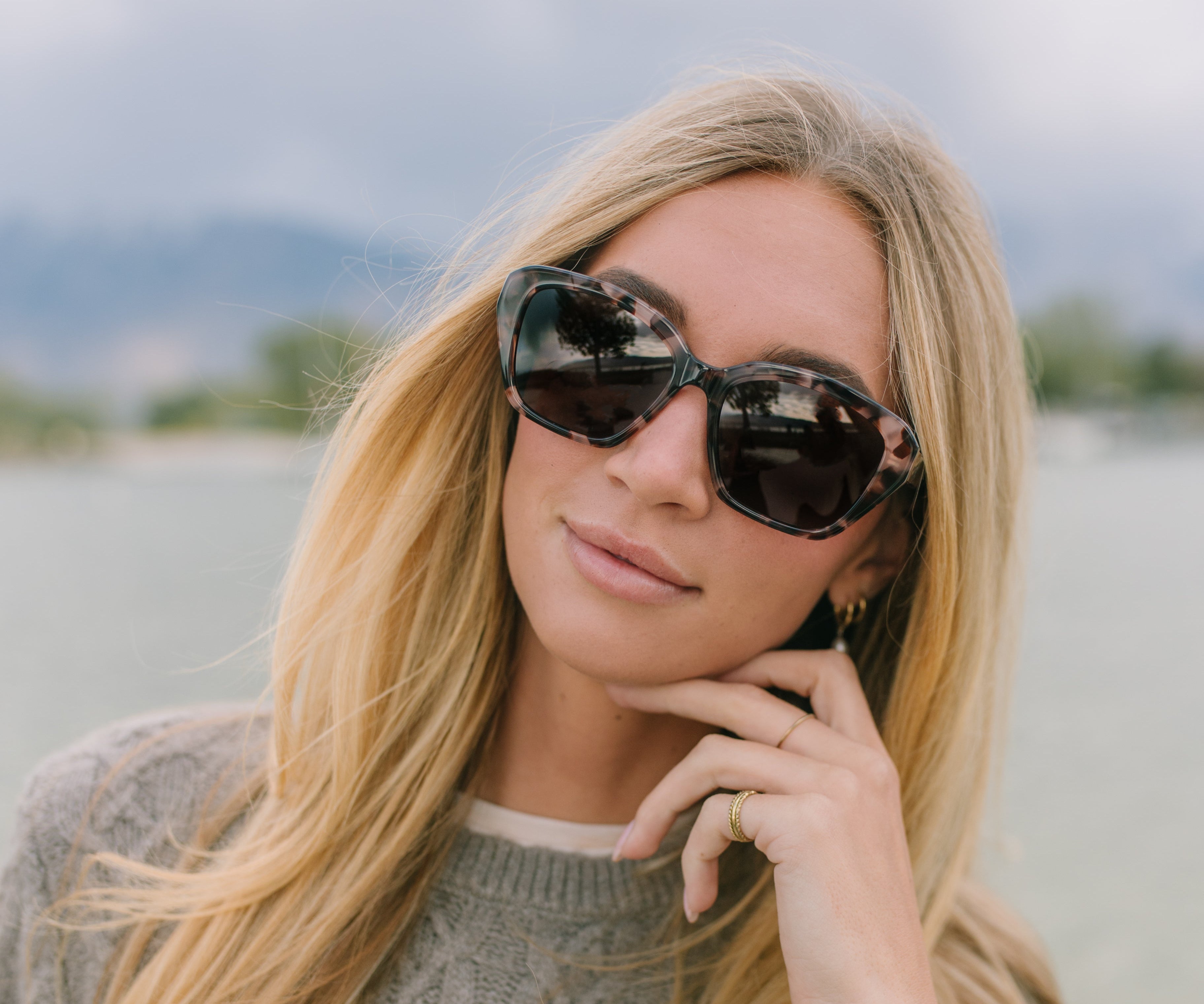 Woman wearing sunglasses and a gray sweater by a body of water with mountains in the background