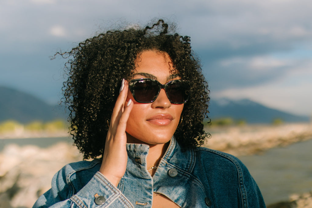 Woman wearing sunglasses and a denim jacket by a body of water with mountains in the background